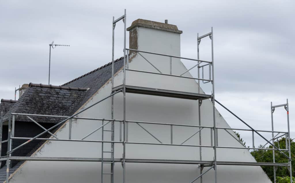 Échafaudage Façade Maison Ardoise Travaux Échafaudage métallique devant le pignon blanc d'une maison avec toiture en ardoise sous un ciel gris et nuageux.