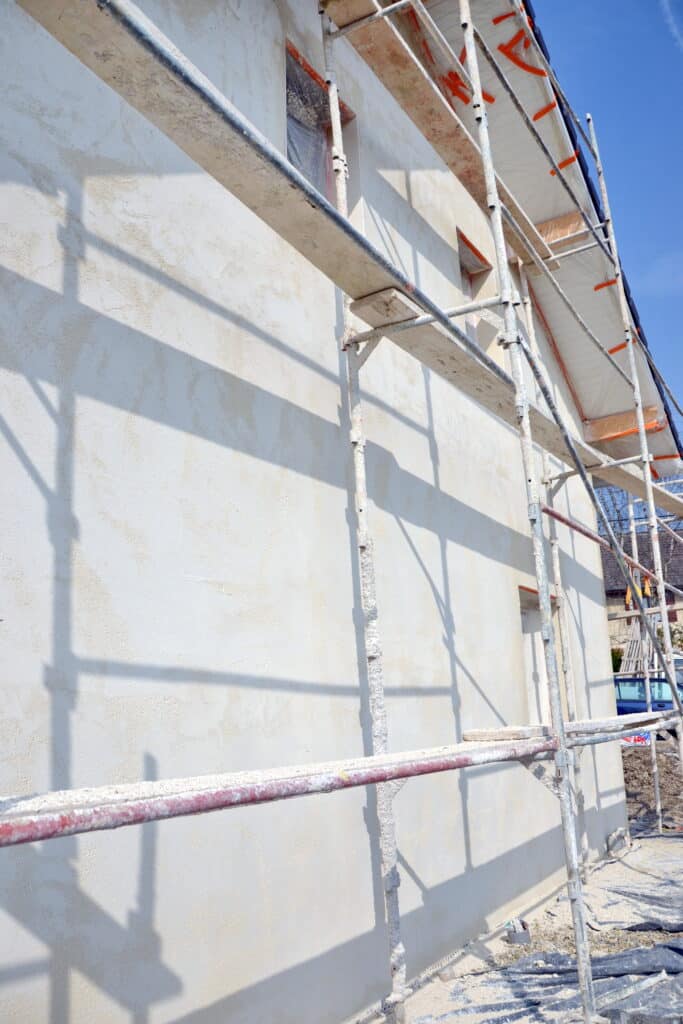 Construction Maison : Enduit Façade et Échafaudage Façade d'une maison neuve avec enduit clair et échafaudage métallique, sous un ciel bleu clair.