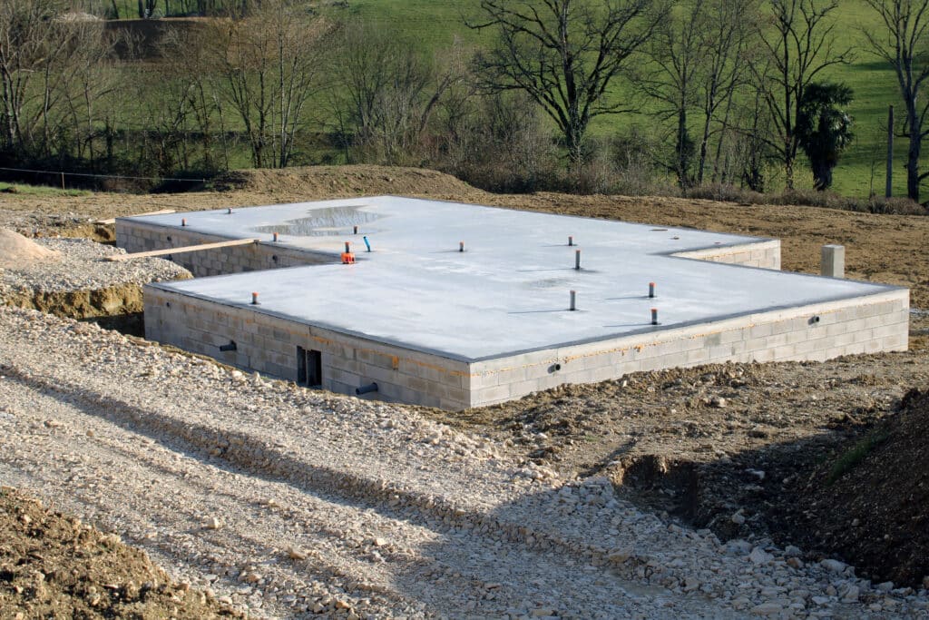 Fondations en parpaings d'une maison en construction, avec dalle de béton fraîchement coulée et tuyaux apparents, chantier rural en arrière-plan.