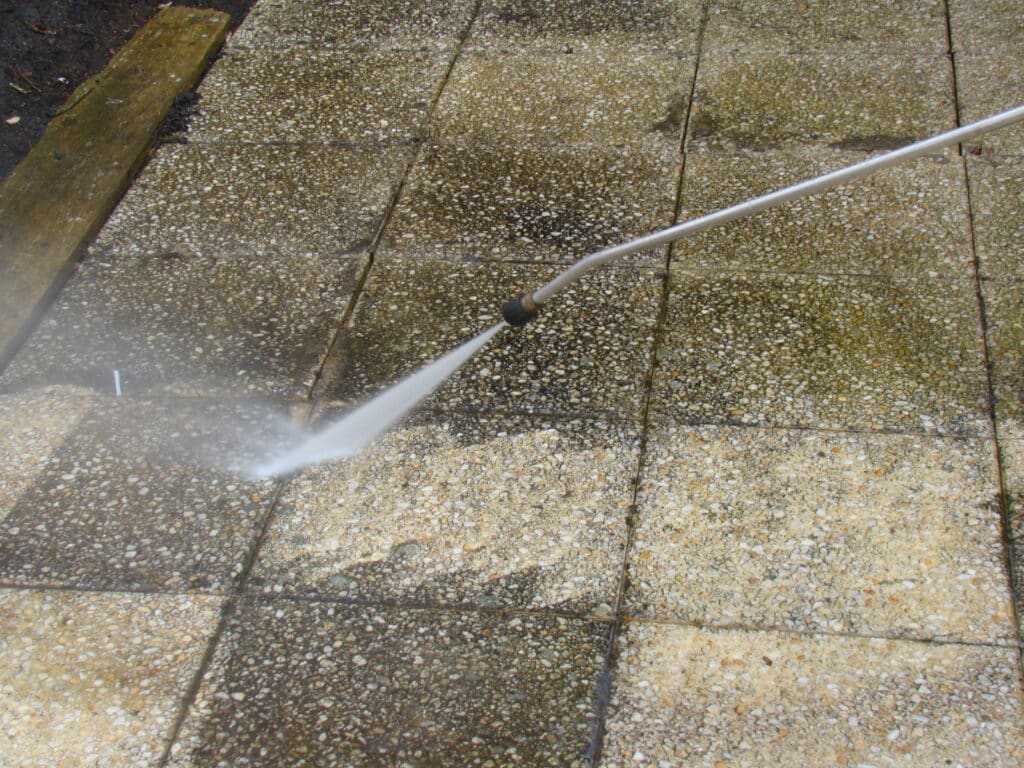 Nettoyage haute pression d'une terrasse en dalles de gravier exposées, révélant la couleur claire des agrégats sous le jet d'eau.