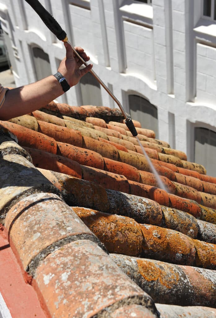 Main lavant des tuiles de terre cuite recouvertes de lichen orange et gris avec un nettoyeur haute pression, devant un bâtiment blanc.