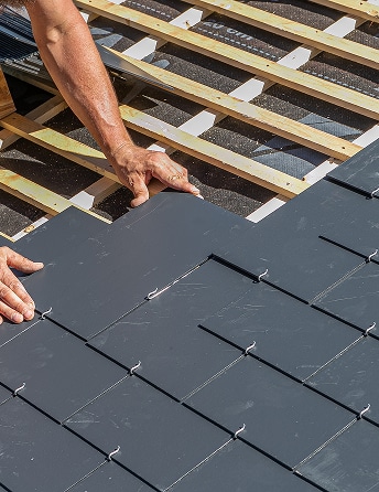 Un couvreur installe des ardoises sombres sur une toiture en construction, fixées sur des liteaux en bois et une sous-couche visible.