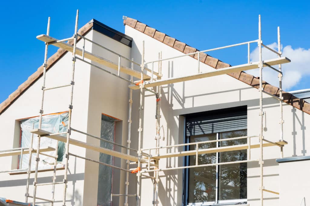 Échafaudage devant la façade crème d'une maison en construction ou rénovation, avec tuiles de toit visibles et fenêtres protégées.