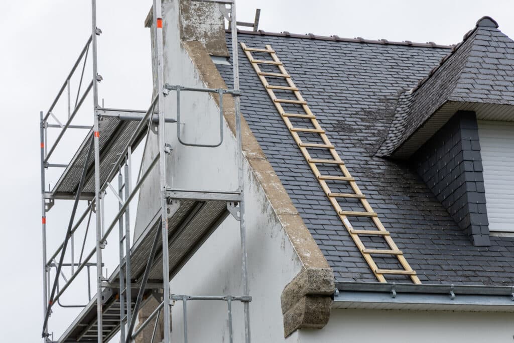 Rénovation Toiture Ardoise : Échafaudage et Échelle de Secours Échafaudage métallique contre une façade blanche avec cheminée, une échelle de toit en bois sur un toit en ardoises sombres en cours de rénovation.