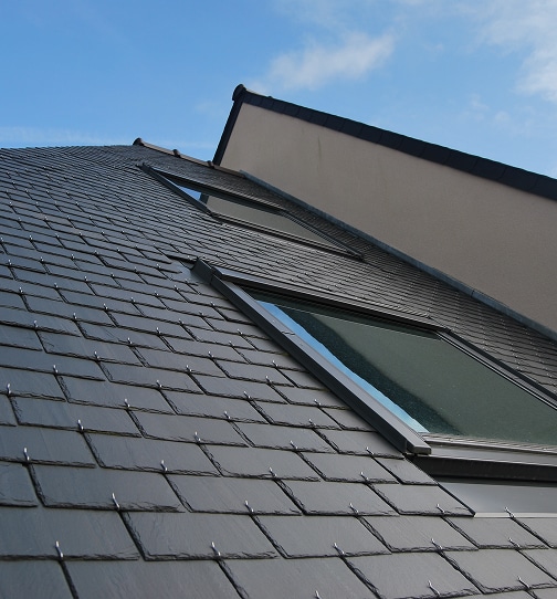 Toiture en ardoises grises avec deux fenêtres de toit (Velux) intégrées sous un ciel bleu clair, contrastant avec un mur de façade beige.
