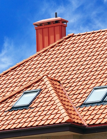 Toit en tuiles terre cuite, avec une cheminée rouge et deux fenêtres de toit (velux) sous un ciel bleu vif.