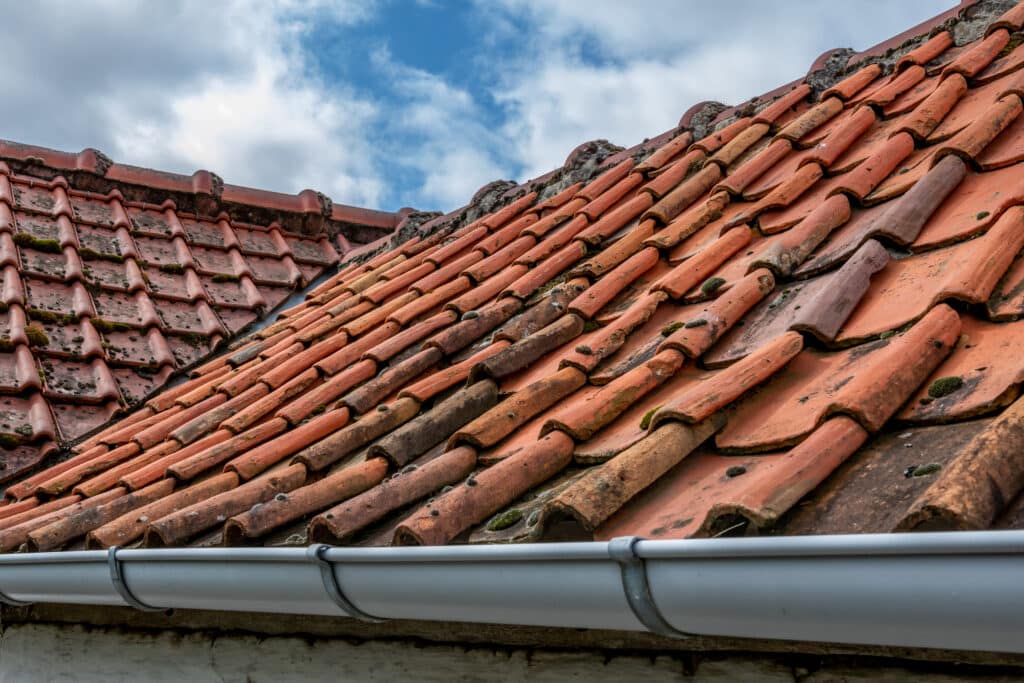Toiture ancienne en tuiles romanes rouges, partiellement couvertes de mousse, avec une gouttière grise neuve sous un ciel nuageux.